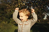 A toddler with curly blonde hair holds his arms above his head in enthusiasm