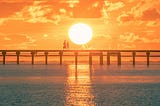 Two silhouettes stand at the end of a pier, bathed in the warm orange glow of a setting sun.