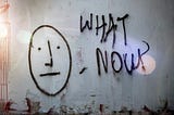 Greyish concrete wall of what appears to be an abandoned building with “What Now?” scrawled haphazardly next to a simply drawn face with a straight mouth.