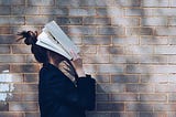 Backdrop: a brick wall. A young woman with a high bun wearing a black blazer holds a book over their face.