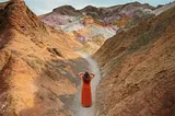A lady in a long red dress, touching her hair, walks away from the camera on a curving trail between two steep, colorful red clay inclines.
