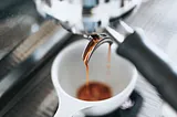 This is a photo of an espresso shot being pulled into a white cup. The coffee is flowing in a steady stream, indicative of a well-extracted espresso, and you can see the crema forming on top, which is the golden frothy layer that is often a sign of good quality espresso. The background is blurred, focusing the attention on the coffee extraction process.
