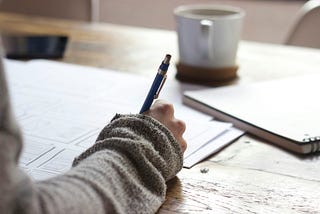 A person writing on paper with a mug and notebook off to the side.