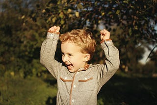 A toddler with curly blonde hair holds his arms above his head in enthusiasm