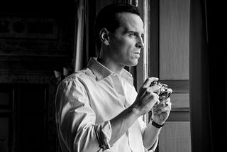 Photo shows a 47-year-old version of Tom Ripley, played by Andrew Scott, apparently wearing a wig and staring out a window in a crisp white cotton shirt, posing with an old 35mm camera.