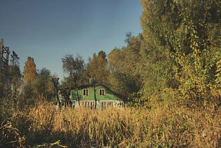 A field with grass with a falling apart house behind. Forest surrounds the house.