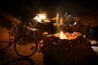 A double exposure showing a bearded man sitting at a campfire looking at a lantern, and reading a book by headlamp