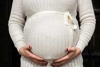 Close-up of pregnant torso in a white sweater holding their baby bump. When a woman is five months pregnant with a baby girl, the cellular beginnings of all of the fetus’s eggs have developed.