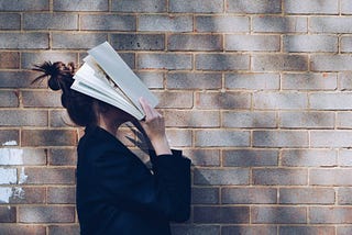 Backdrop: a brick wall. A young woman with a high bun wearing a black blazer holds a book over their face.