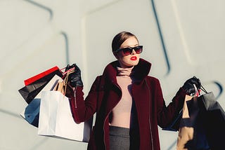 A chic woman in sunglasses holds many shopping bags. The light shines on her face, the background is white with a triangular raised design.