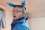 An Asian fellow wearing a blue puffy jacket, and a headband with two long blue ears and dangling blue ribbons.