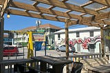 Wood pergola over picnic tables and chairs. A yellow sign — “Island Bar” with a list of a few cocktails. A colonial-style building is at the back of the parking lot with another plain white two-story building next to it. There is pink and grey street art on the white building, that says “Heel 2 Toe”