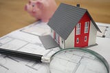 A house with a piggy bank in the background and a magnifying glass in the foreground on top of blueprints.