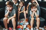 five kids sit in the back seat of a van, all looking sad. snacks are strewn about the van.