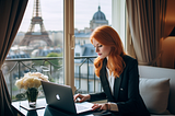 Woman contently sitting in hotel room in Paris working at laptop next to window with Eiffel Tower in the distance