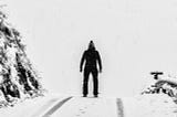 A person wearing all black clothing stands atop a snowy hill.