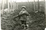 A black and white photo of a young boy of about four, the author, who stands between two barbwire fences at an Eastern European border. The boy is on the muddy pass and holding a large AK 47 gun with an afixed bayonet.