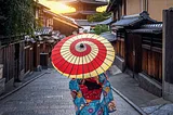 A Japanese woman walks down a glowing Kyoto street with a red and cream parasol over her shoulder.
