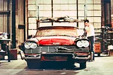 A red 1958 Plymouth Fury car which has a cracked windshield and various dents and broken body panels. There is a man getting into the car. It is a scene from the movie, Christine.