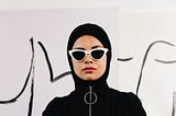 The image shows a woman wearing a black hijab, tucked into a O-ring zip sweatshirt, and white cat-eye sunglasses.