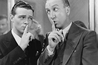 Black and white photo of two men with fingers to the lips: “ssssh”