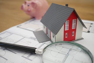 A house with a piggy bank in the background and a magnifying glass in the foreground on top of blueprints.