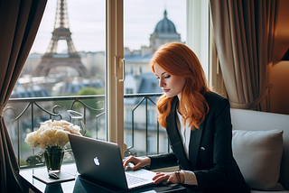 Woman contently sitting in hotel room in Paris working at laptop next to window with Eiffel Tower in the distance