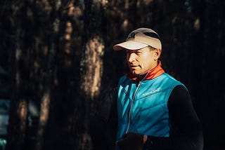 Author, a smiling middle-aged man, highlighted by morning sun wearing a ball cap, a blue runner’s vest, black long-sleeved shirt and black gloves.