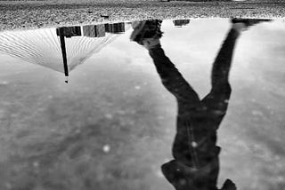 a black-and-white photo showing a person running and a bridge reflected in a puddle of water on asphalt