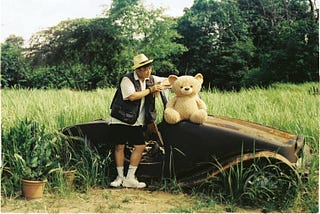 A man in a hat stands next to a teddy bear by a car wreckage on the grassland