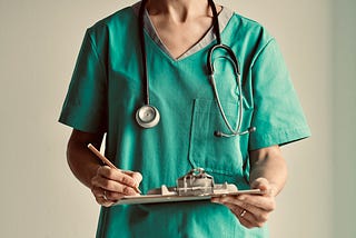A young woman in green hospital scrubs and stethoscope holding a clipboard and pen