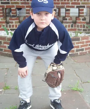 Little boy in baseball uniform, holding his glove while squatting down and looking into the camera