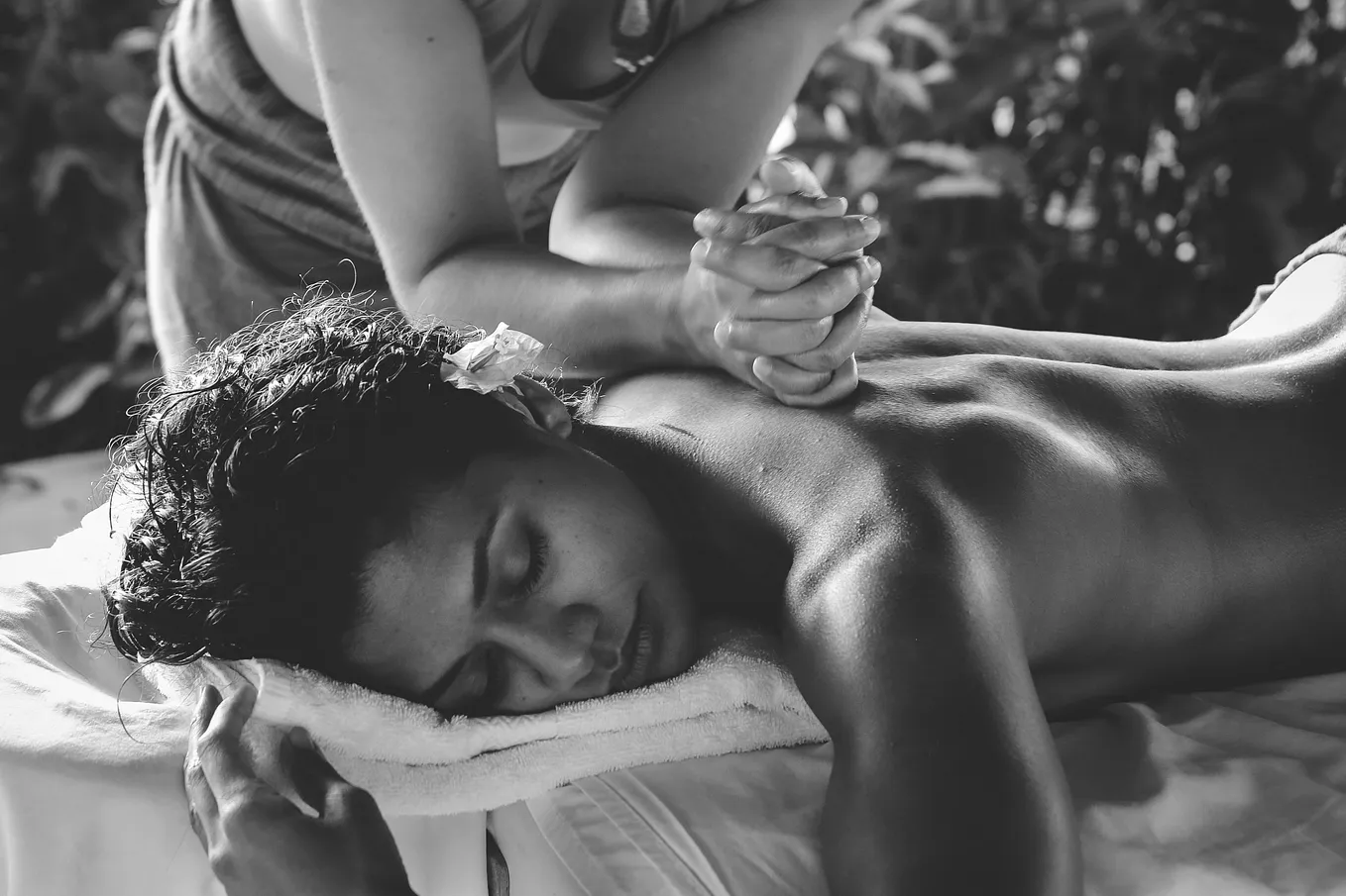 A woman lies on a bed being given a massage, her eyes closed, a flower behind her ear. The scene is one of tropical luxury