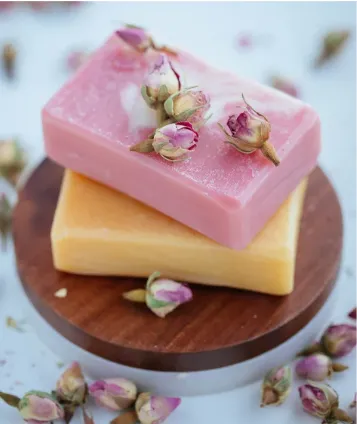 A photograph of pink and light orange bars of soap stacked together displayed on a round wood grained container. There is a sprinkling of flower buds on top of the pink bar of soap and around the container. The surface and the blurred background are light blue.