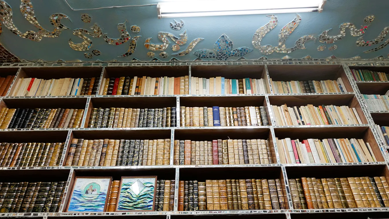 A curated bookshelf with classically bound tomes against a light blue ceiling decorated with mirror mosaics