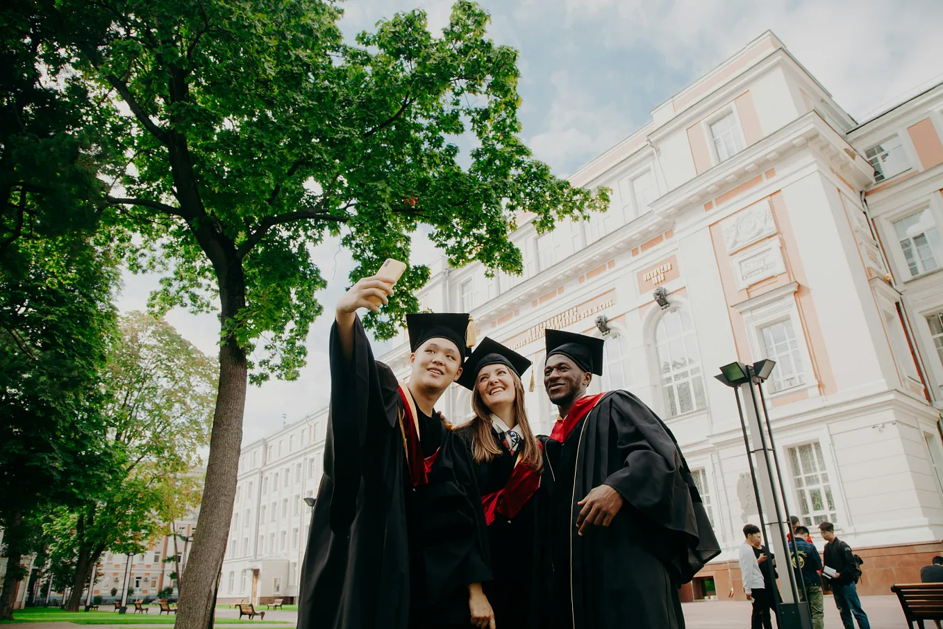 Three college graduates in commencement robes taking a photo outside of a university building