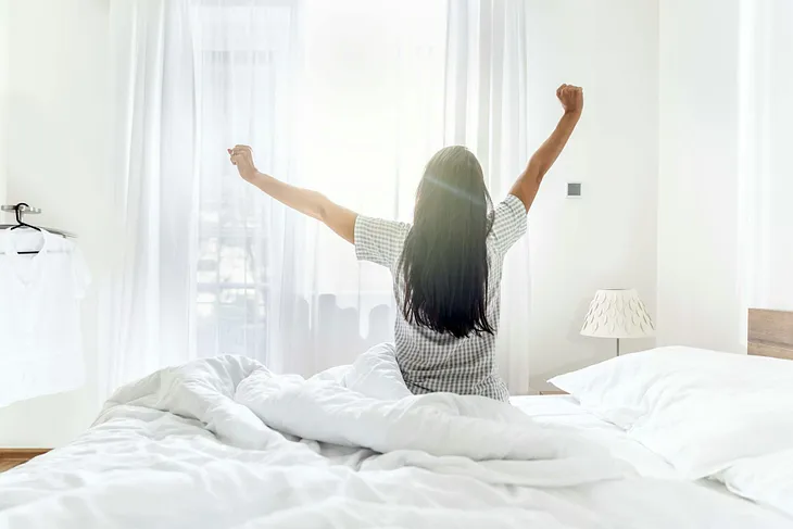 A woman sitting up in bed and stretching as the morning light streams in through a window