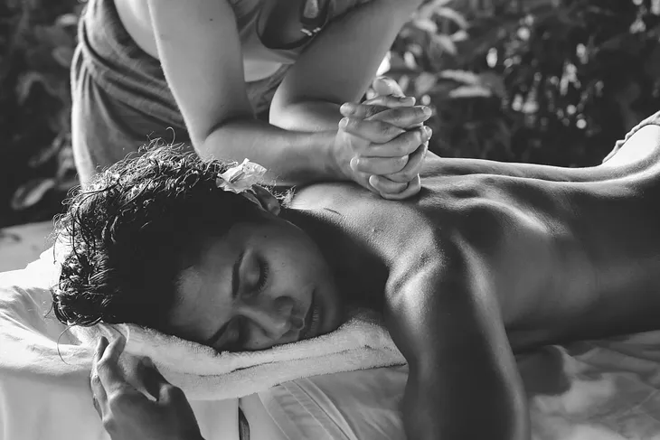 A woman lies on a bed being given a massage, her eyes closed, a flower behind her ear. The scene is one of tropical luxury