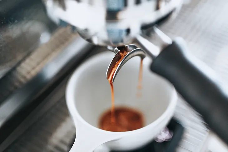 This is a photo of an espresso shot being pulled into a white cup. The coffee is flowing in a steady stream, indicative of a well-extracted espresso, and you can see the crema forming on top, which is the golden frothy layer that is often a sign of good quality espresso. The background is blurred, focusing the attention on the coffee extraction process.