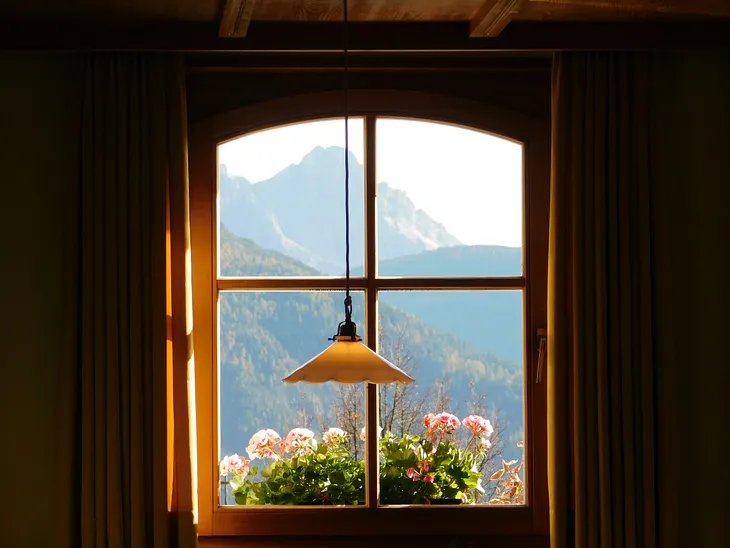 A lamp hanging in front of a window, with a flower box and mountain visible outside the window.