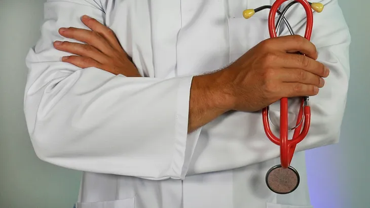 Arms folded over chest, male hands, wearing white lab coat, right hand holds a folded red stethoscope, a doctor.