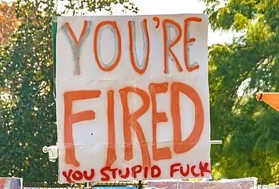 White protest sign that says in large orange letters “You’re fired” with much smaller red letters that say “you stupid fuck” below. Trees in the background.