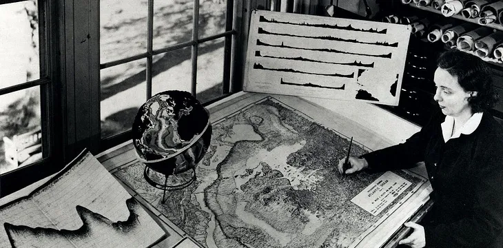 Black and white photo of a woman writing on a large map of part of the seafloor that covers the entire desk, with a globe sitting on it