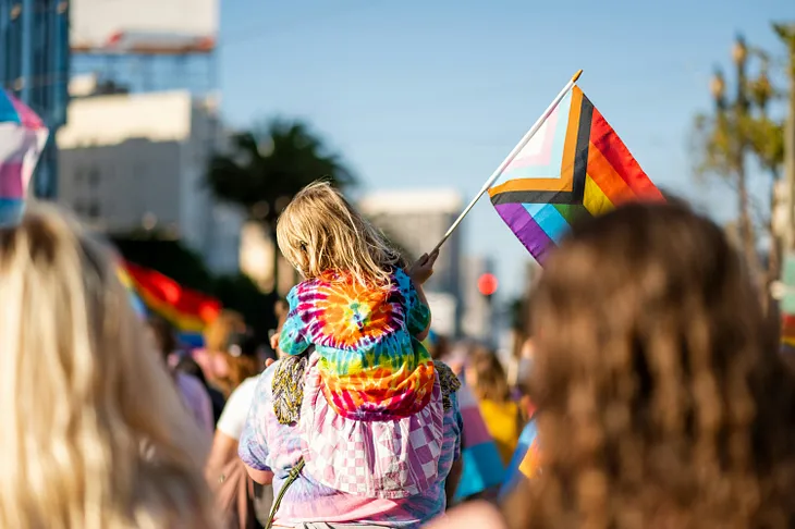 A young child wearing a tie-dye shirt and holding the Pride flag sits atop their parent’s shoulders at a Pride parade