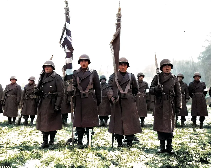 Two color guards and color bearers of the Japanese-American 442d Combat Team, stand at attention, while their citations are read. They are standing on ground in the Bruyeres area, France, where many of their comrades fell.