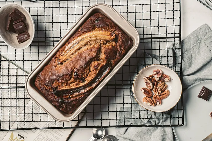 Image shows banana bread in a tin, in the middle of the image, then a bowl with pieces of chocolate in the top left corner and a bowl of pecan nuts in the bottom right corner. All of this are on a black metal rack.